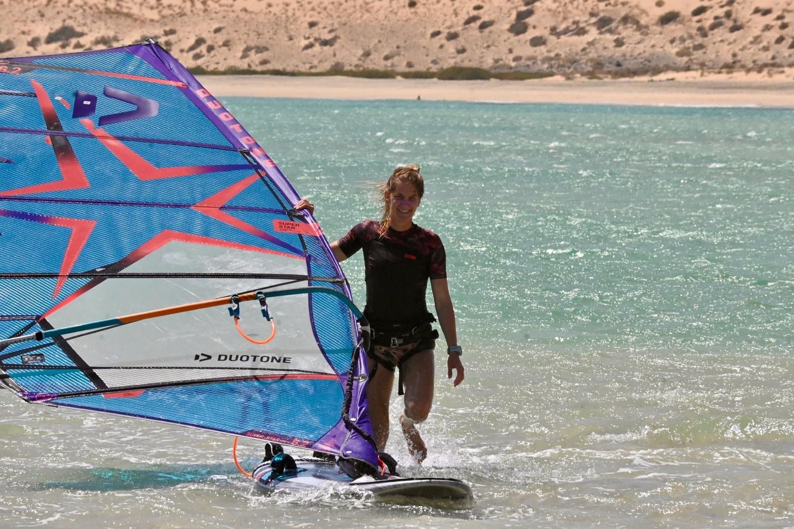 Person windsurfing on turquoise water near a sandy beach, holding a colorful sail.