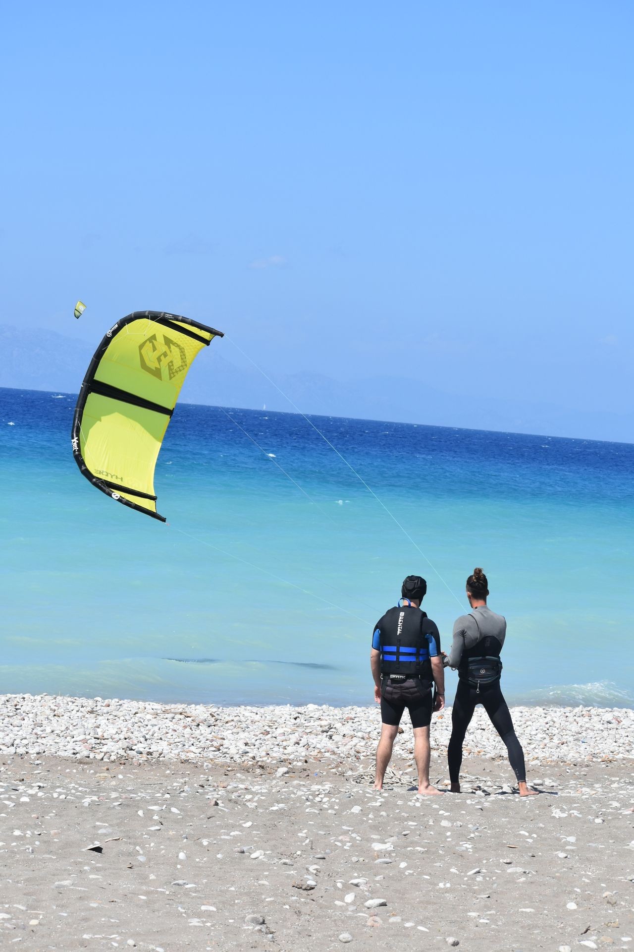 Two people on a beach watching a yellow kite soar over the ocean.