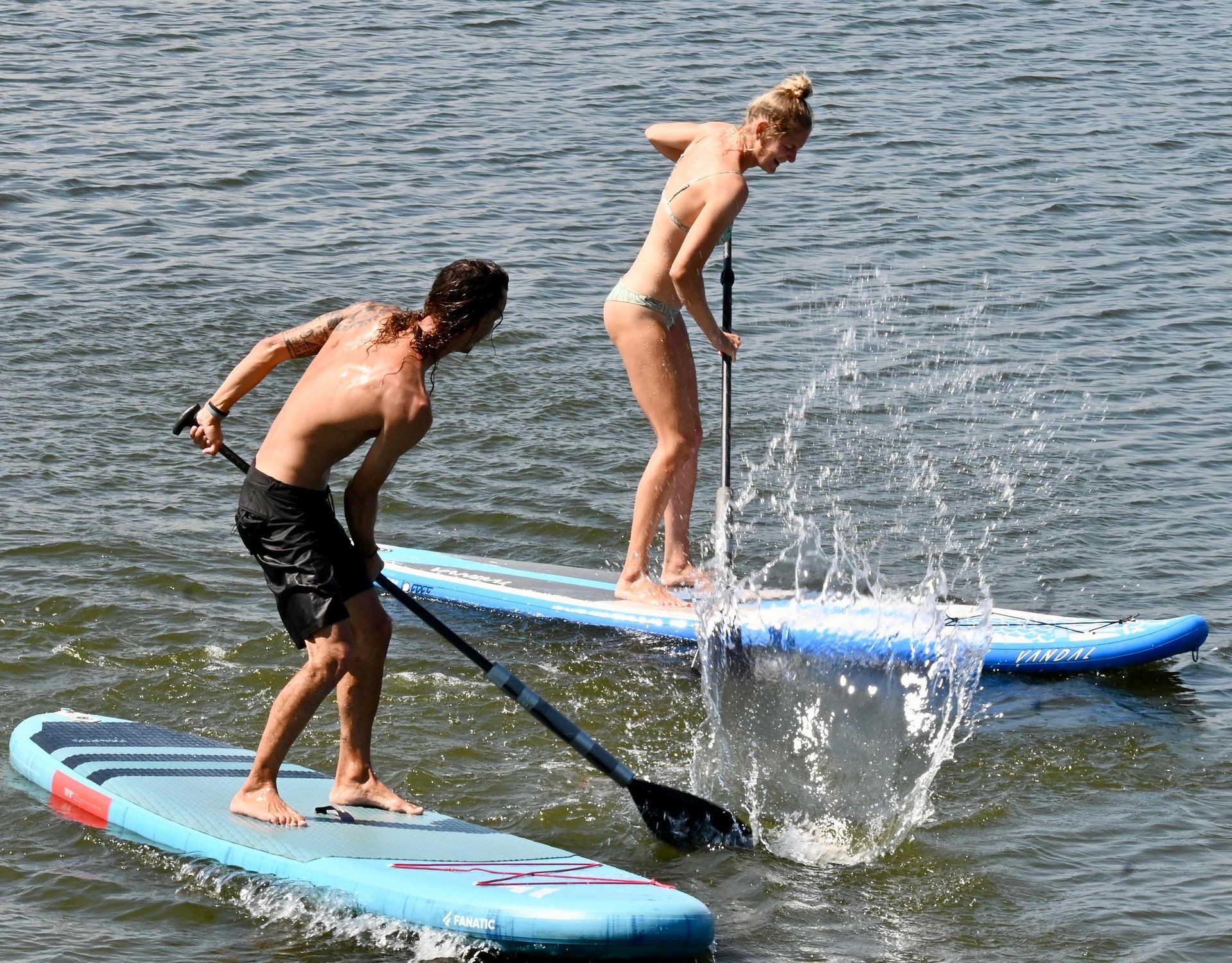 Two people paddleboarding on a sunny day, splashing water as they move across the surface.