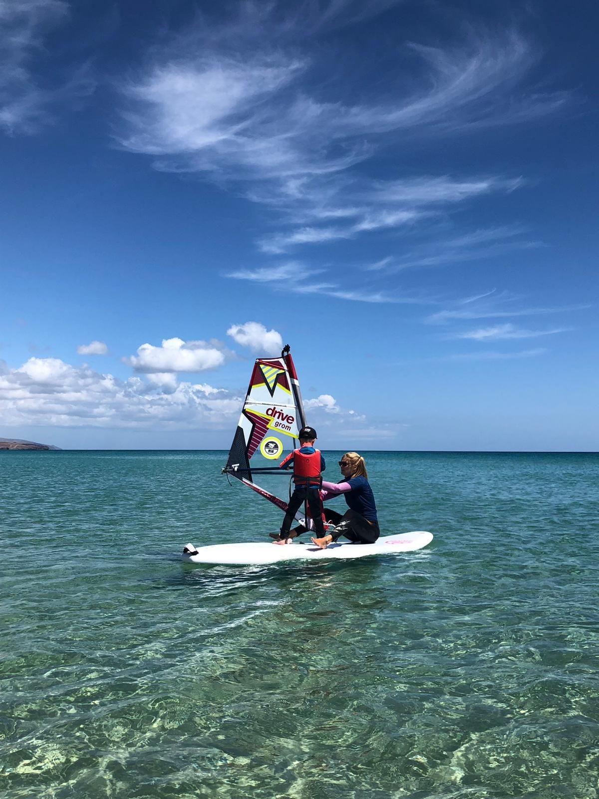 Two people windsurfing on a clear blue sea under a bright sky with wispy clouds.