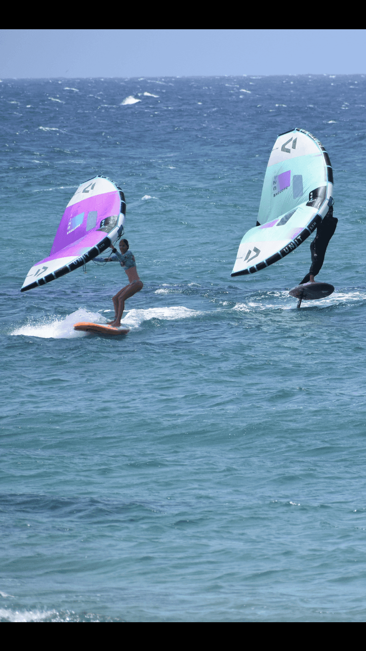 Two people wing foiling on blue ocean waves with colorful wings.